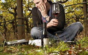 Gary Vaynerchuk holding a glass of Eric Ross Pinot Noir sitting cross-legged in the woods with the wine bottle and laptop